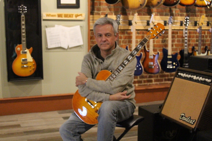 Chris Mitchell holding a CMG guitar next to a Devilcat amplifier inside of his store in downtown Statesboro.