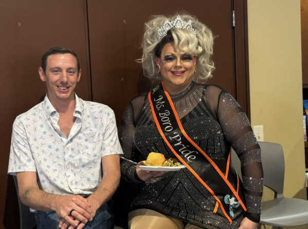 Man on left sits with drag queen on right holding a plate of food.
